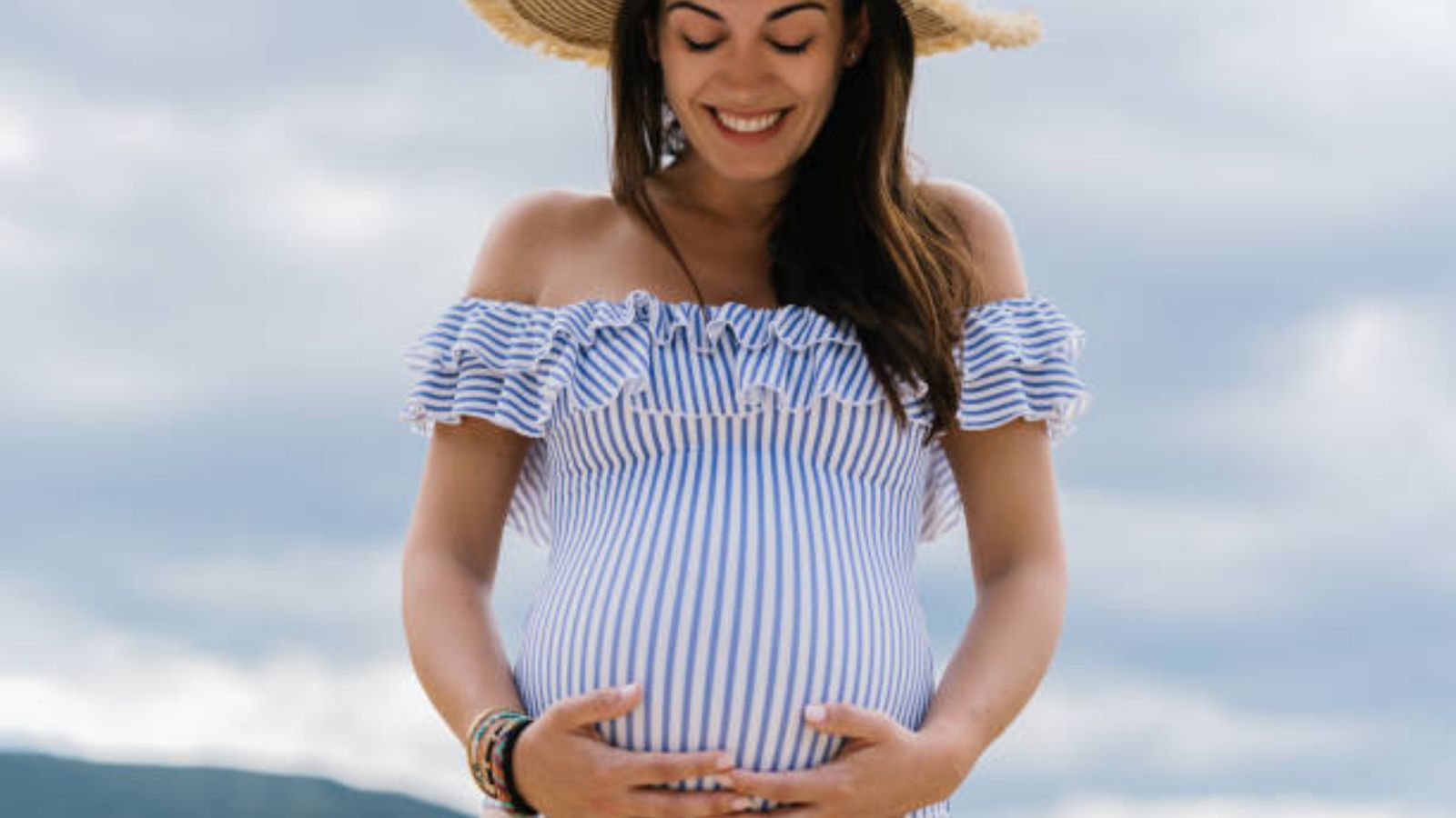 A smiling pregnant mother in a striped dress, hands on her bump, under a bright sky